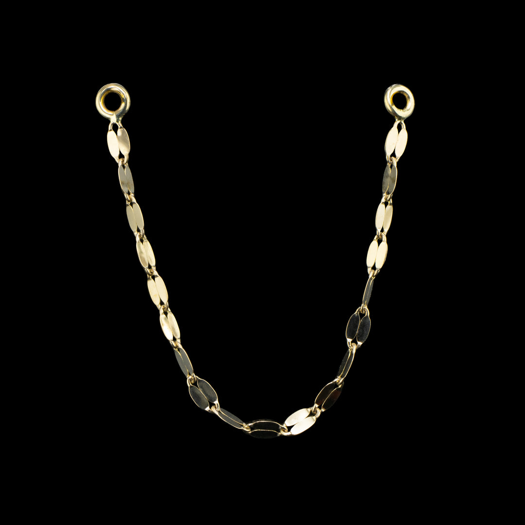 A single yellow gold tile chain on a black background. a tile chain consists of flattened links that reflect light. two jump rings on either side for conch, nostril, helix, cartilage piercings.