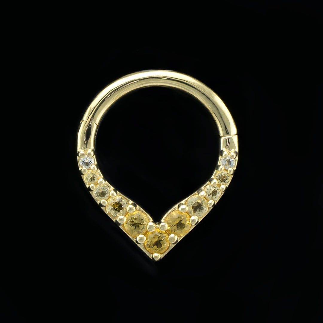 A yellow gold piece of body jewelry on a black background. This is a hinge style ring. The design consists of a solid gold ring, rounded at the top and coming to a point at the bottom, creating a V shape. There are eleven gemstones set in prongs along the V shape with the largest stone at the center of the point and descending sizes. The stones start with a darker citrine at the center and get lighter as they go up the sides of the V.