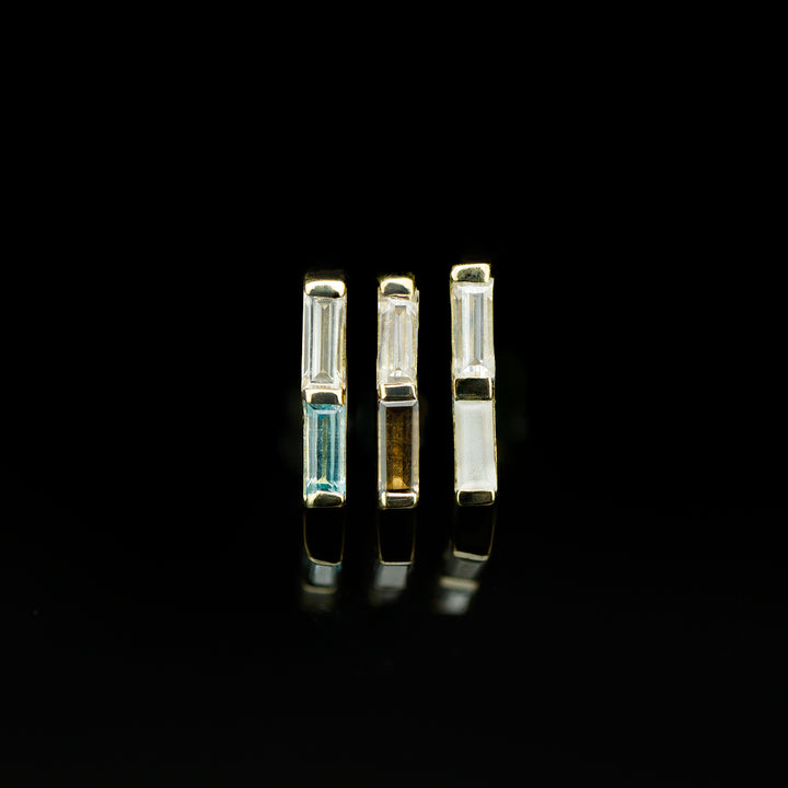 Three bar shaped pieces of body jewelry with two rectangle shaped gemstones set in channels on each. The piece of the left feature a white cz set atop light aquamarine, the middle is a white cz atop brown Smokey quartz, and the last a white cz set atop a sandblasted white cz. The sandblasting makes the stone look opaque and matte.  