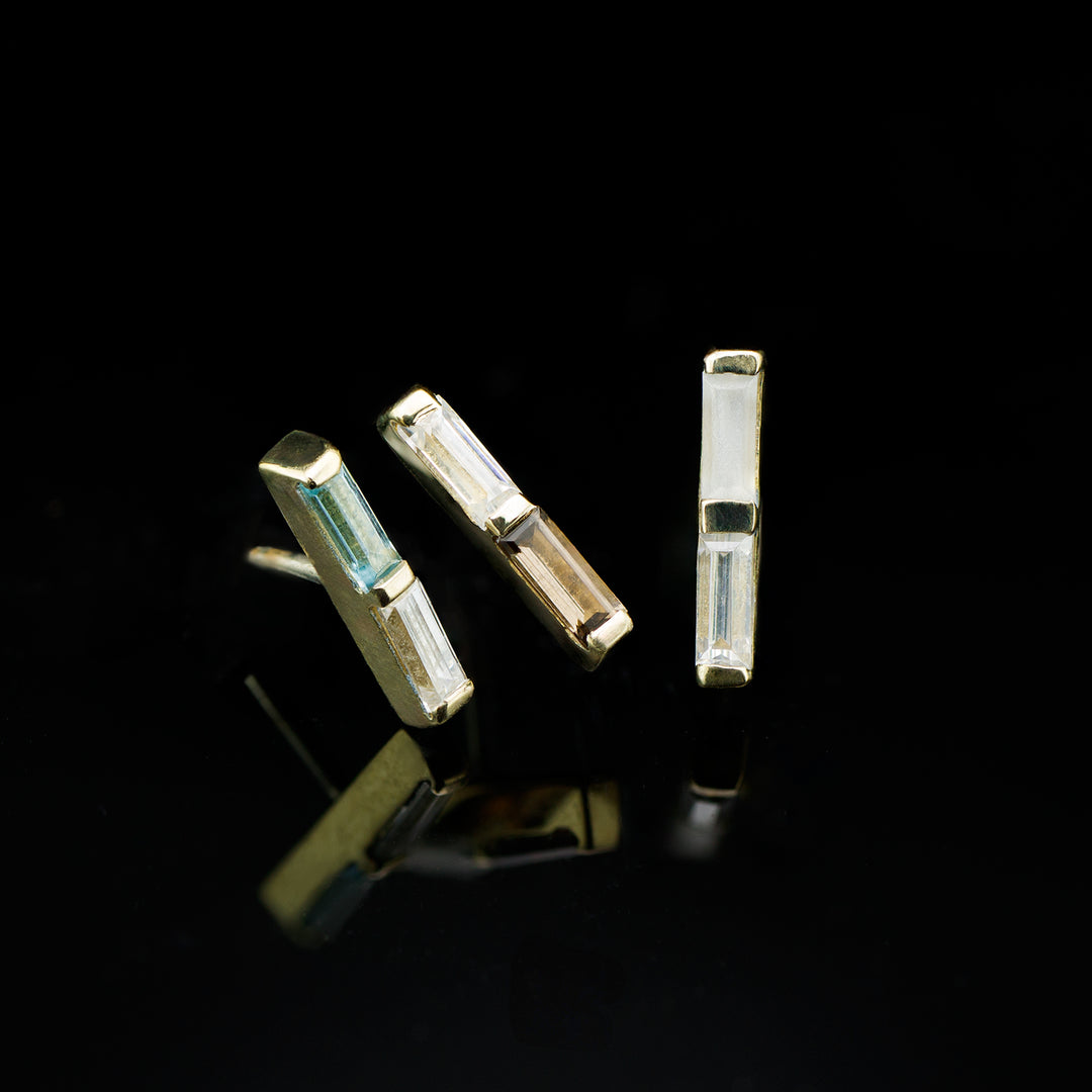 Three yellow gold bar shaped pieces of jewelry with two gemstones set in each. Arranged on a black background. 