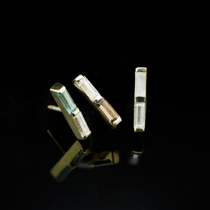 Three yellow gold bar shaped pieces of jewelry with two gemstones set in each. Arranged on a black background. 