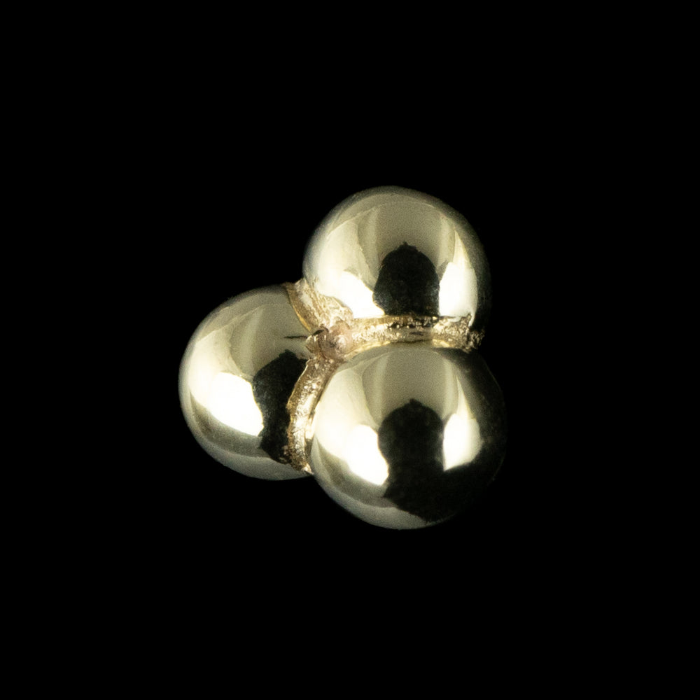 A piece of solid yellow gold body jewelry set on a black background. This photo offers a side view of the piece. This design consists of three small balls sawdered together in a triangular shape. 