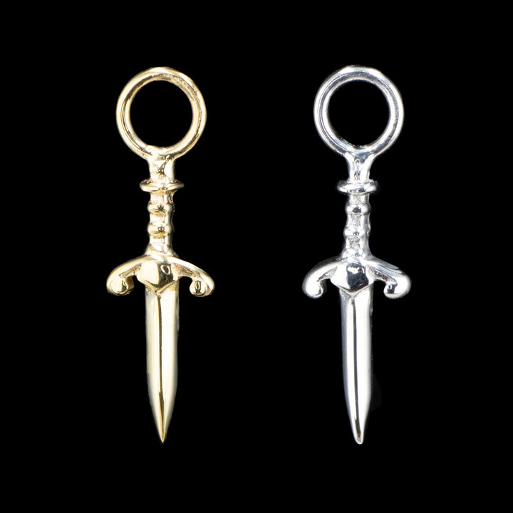 two pieces of fine gold body jewelry on a black background. The left piece in yellow gold and right in white gold. They are dagger design with a decorative hilt and a jump ring at the top. 