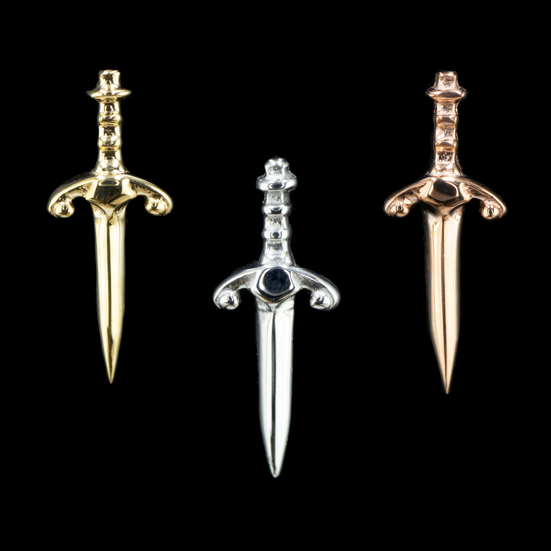 three pieces of body jewelry on a black background. From left to right there is a yellow gold, a white gold, and a rose gold piece. The gold jewelry design is a dagger with a decorative hilt.  