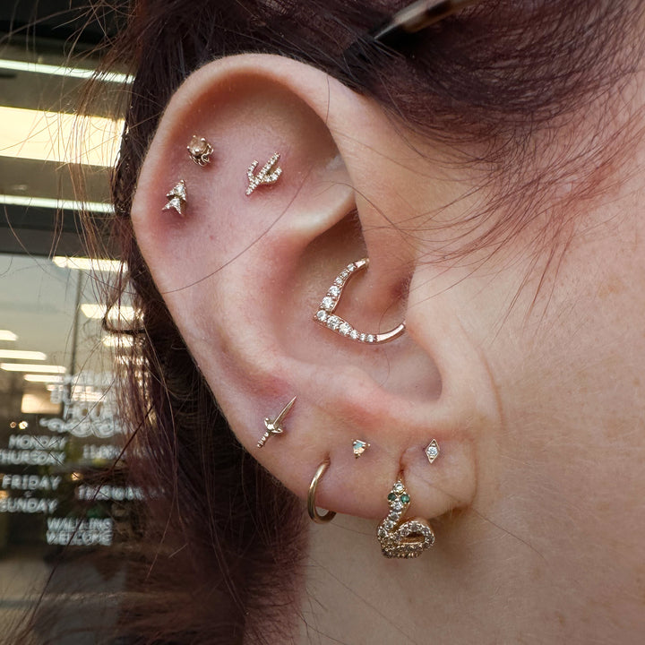 A photo of a clients ear in front of Deep Roots piercing and tattoo. The client is wearing a rose gold Blade end in there third lobe piercing and a Rise and Shine clicker in their daith. The rise and shine clicker is a v shaped ring in rose gold with white cubic zirconia in ascending sizes ending with the largest gem at the center of the point. 