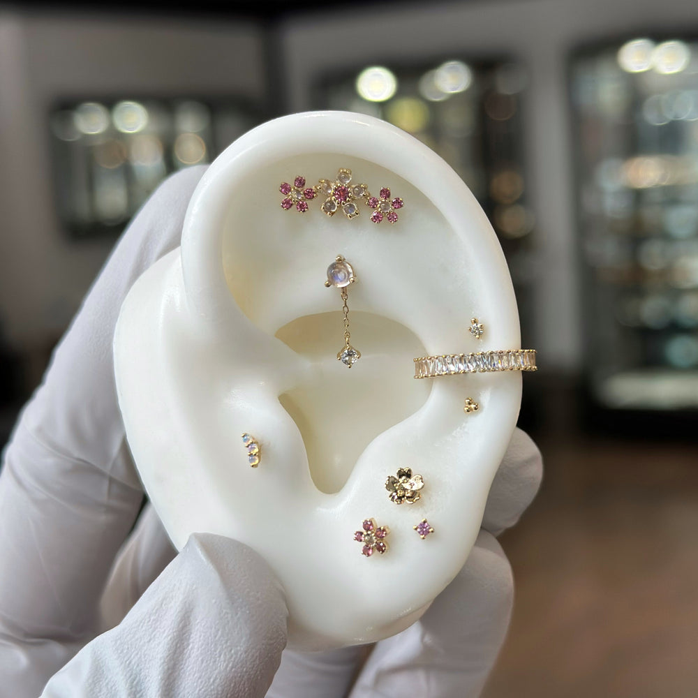 A white silicone ear display with several piercings including a conch with the Cult Favorite Clicker, Flat with the Bianca Threadless End from Buddha, an earlobe with the Simple Daisy End from BVLA, and more! All in yellow gold with pink gems, White CZ, and rainbow moonstone.