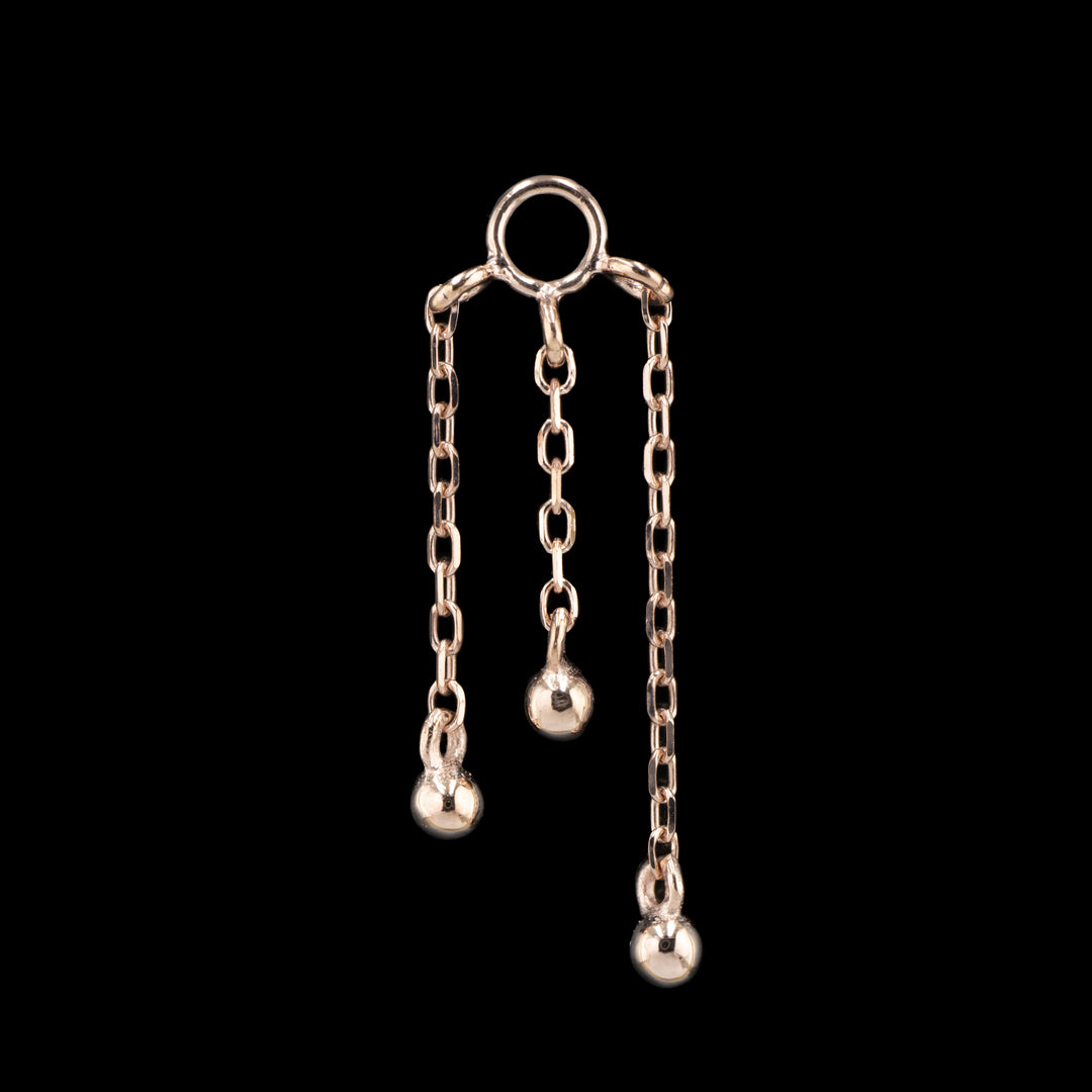 a rose gold piece of body jewelry on a black background. This piece features three varying lengths of cable chain with a small gold bead attached to bottom of each chain.  