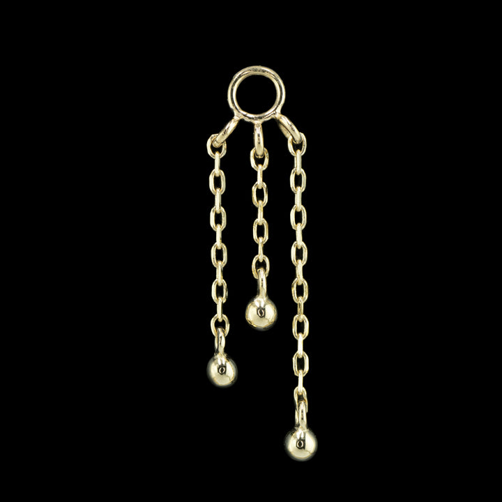 a yellow gold piece of body jewelry on a black background. This piece features three varying lengths of cable chain with a small gold bead attached to bottom of each chain.  