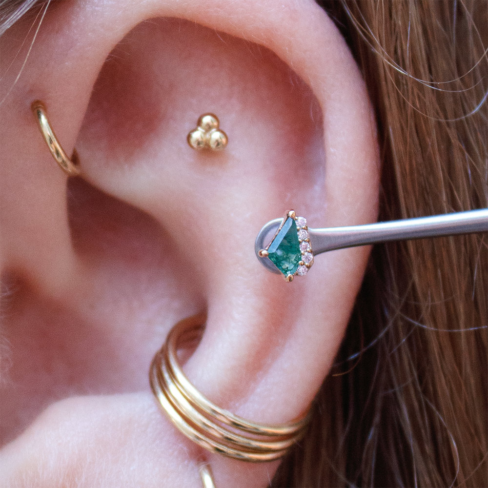 Close up of an ear with multiple piercings including a forward helix, flat and conch piercing. Elevate threadless end with Moss Agate is being held up to the ear in a helix placement. 
Yellow gold jewelry from Buddha Jewelry and Body Vision LA. Cartilage piercings
