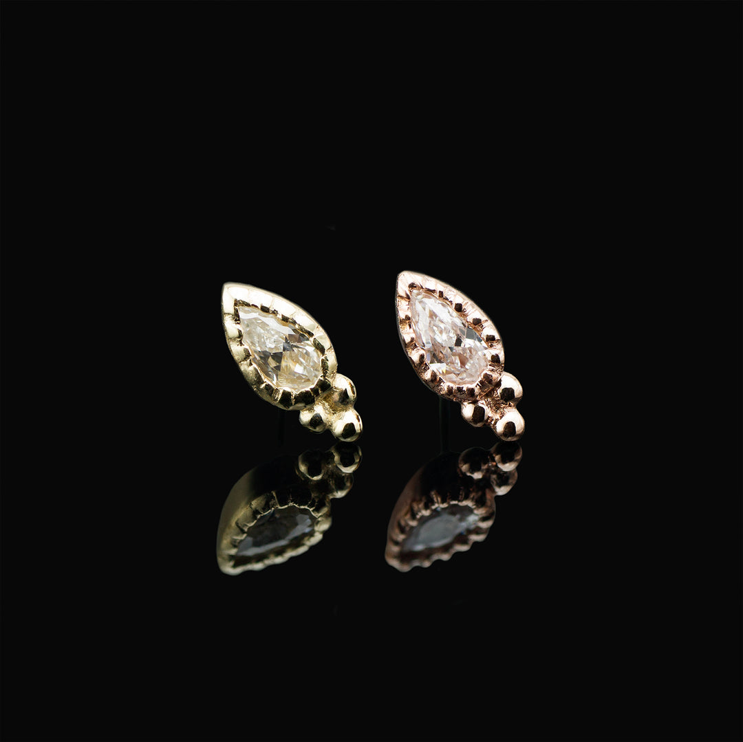 Two gold pieces of body jewelry on a black background. Each have a pear cut white cubic zirconia set in a mill grain bezel with three gold beads arrange in a triangle shape at the bottom of the pear. 