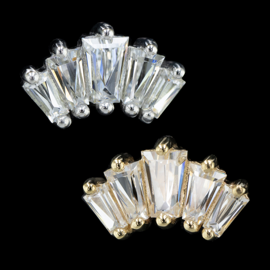 2 pieces of body jewelry on a black background. The top left piece is white gold and the bottom right piece in yellow gold. This piece features 5 tapered baguettes set in prongs. The center cubic zirconia is the largest gem with the outer gems descending in size. 