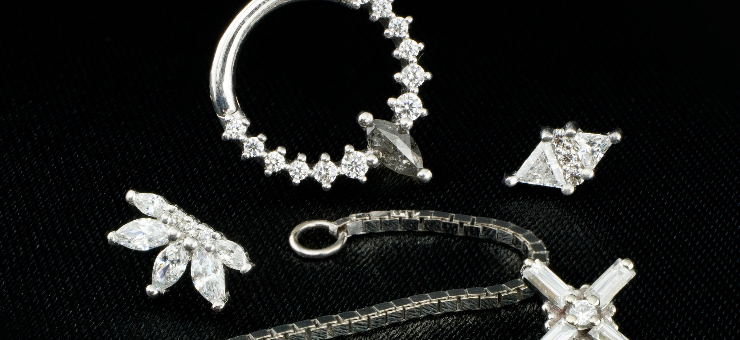 A collection of solid white gold body jewelry pieces on a black cloth background. With white swarovski crystals