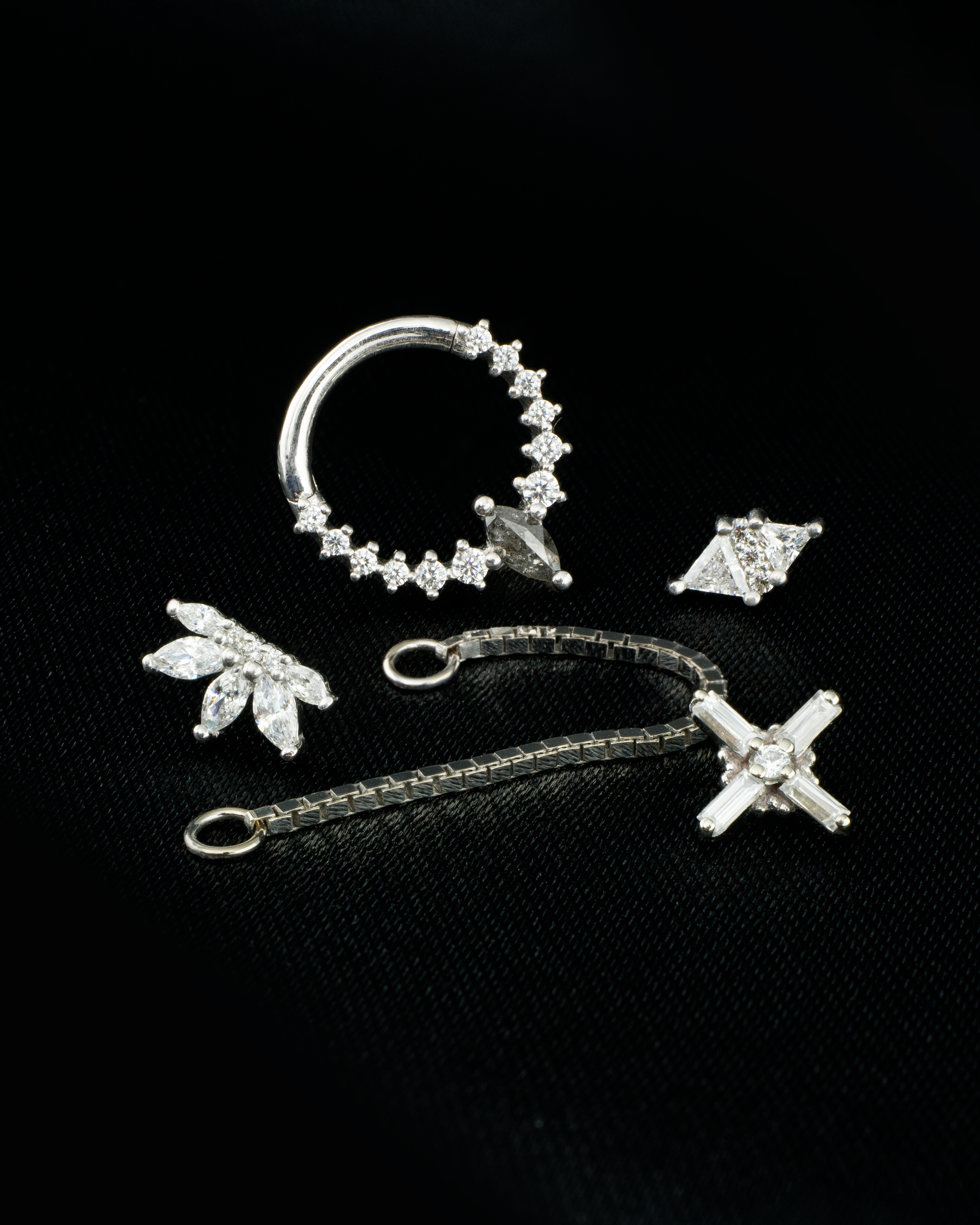 A collection of solid white gold body jewelry pieces on a black cloth background. With white swarovski crystals