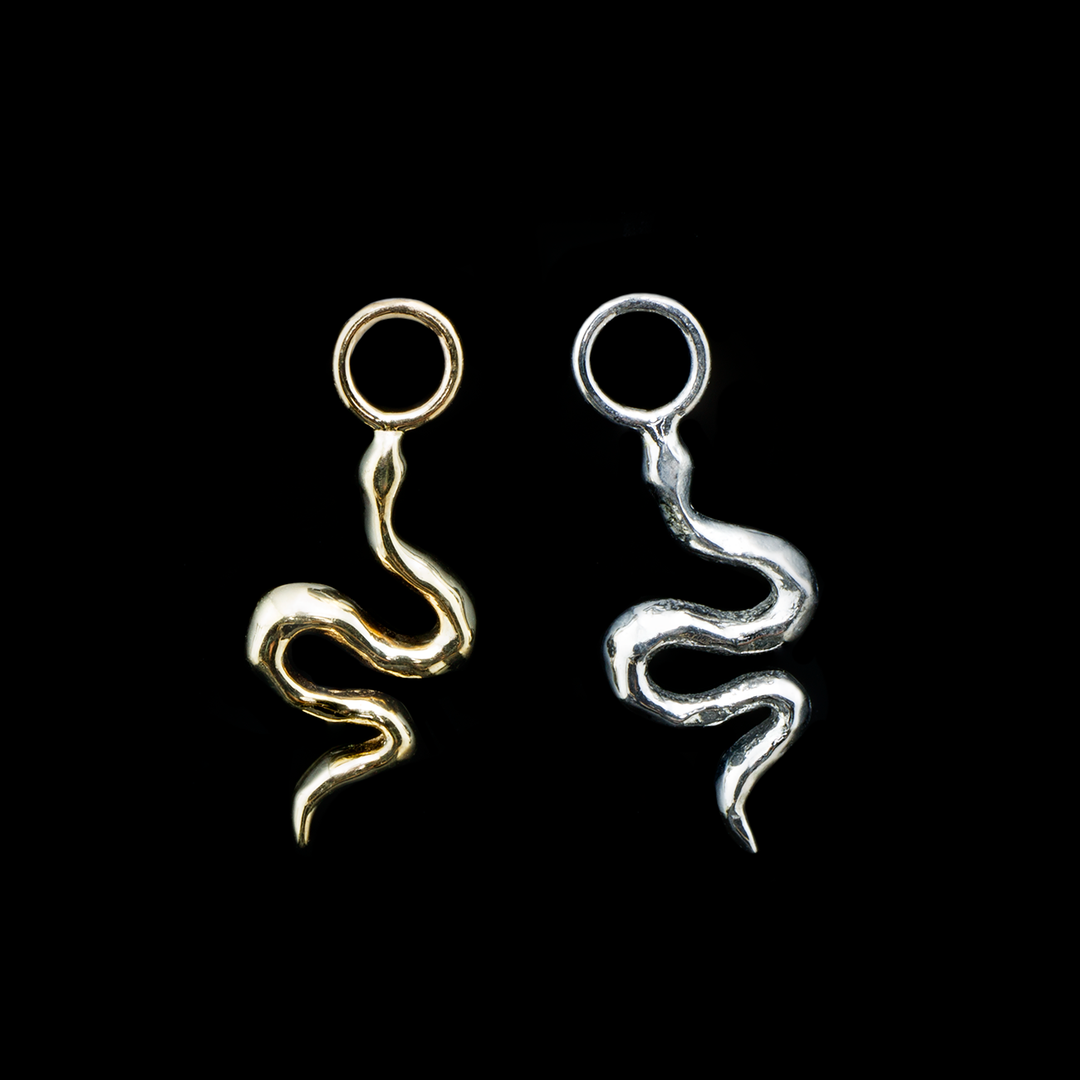 Two Serpent charms on a black background, on in yellow gold and one in white gold. The serpent charm consists of smooth and simple snake shape made of solid gold with a jump ring attached at the head of the snake.