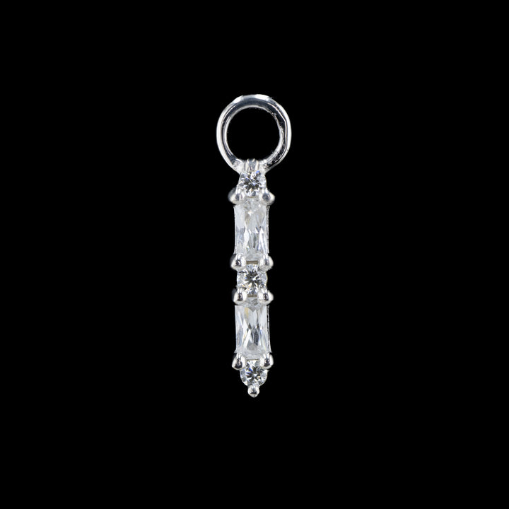 white gold charm on a black background. The piece features 5 swarovski cubic zirconia in a row. There are three round cut white CZs and 2 baguette cut white CZs in an alternating column starting with the round cut cubic zirconia. 