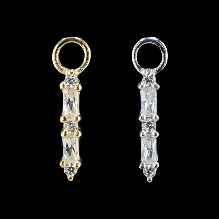 two charms on a black background. The one on the left is yellow gold and the one on the right is white gold. 
The piece features 5 swarovski cubic zirconia in a row. There are three round cut white CZs and 2 baguette cut white CZs in an alternating column starting with the round cut cubic zirconia. 