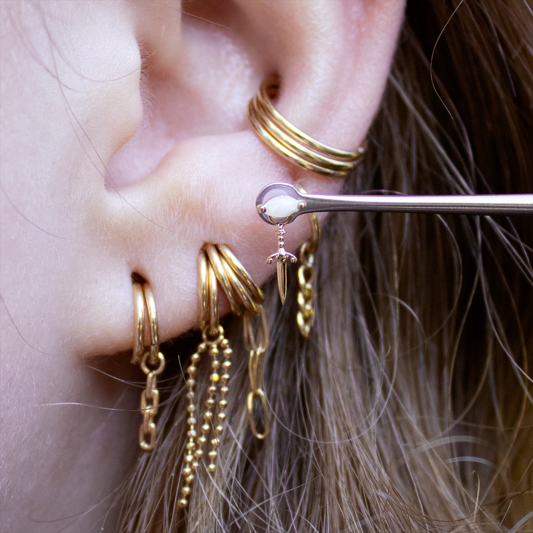 Close-up of the lower half of an ear with multiple earlobe piercings styled with gold seam rings. The white opal zuri and blade charm are held up to the ear showing off an earlobe placement idea. 