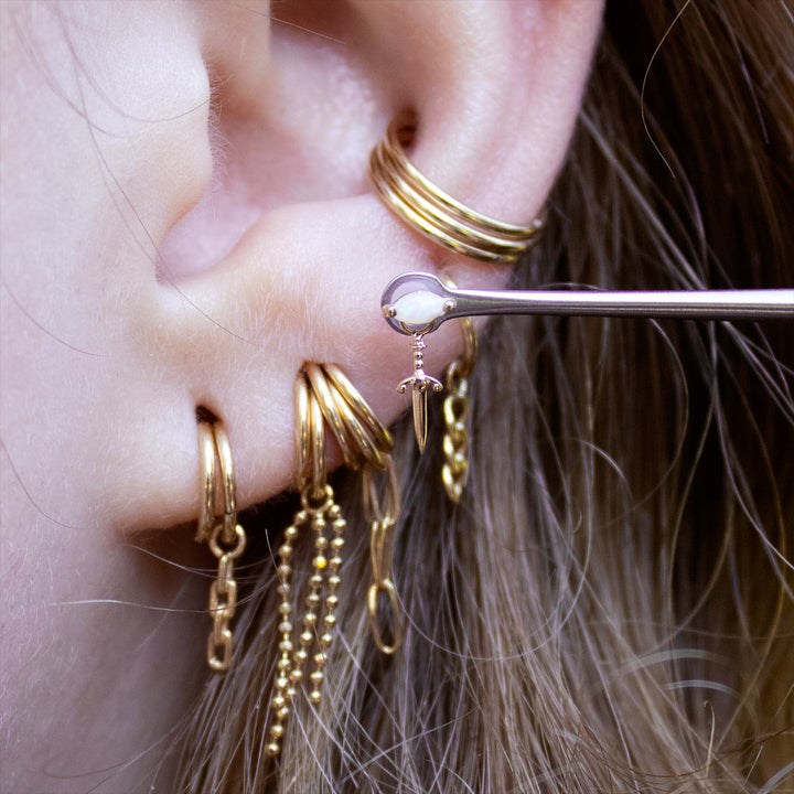 Close-up of the lower half of an ear with multiple earlobe piercings styled with gold seam rings. The white opal zuri and blade charm are held up to the ear showing off an earlobe placement idea. 