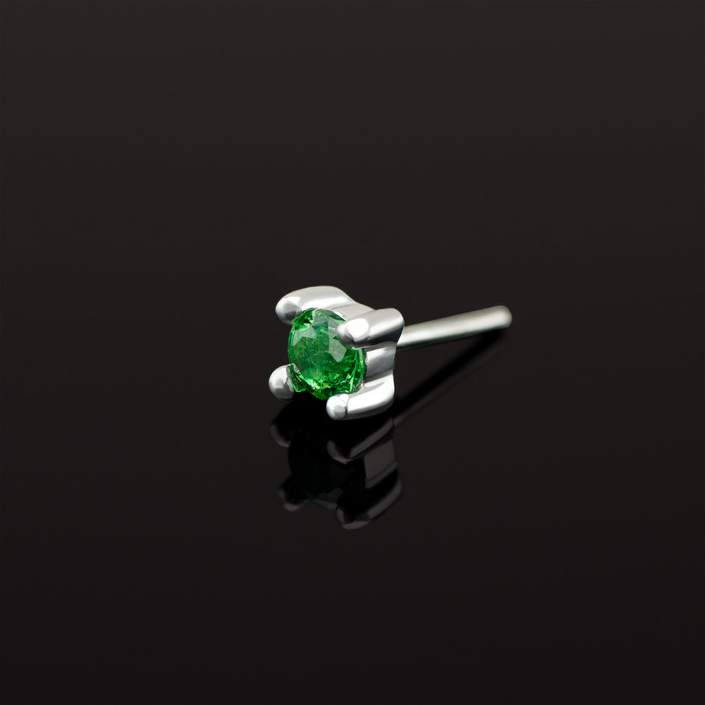 A solitaire brilliant cut green tsavorite set in white gold prongs on a black background. Green garnet end for nose and ear piercings