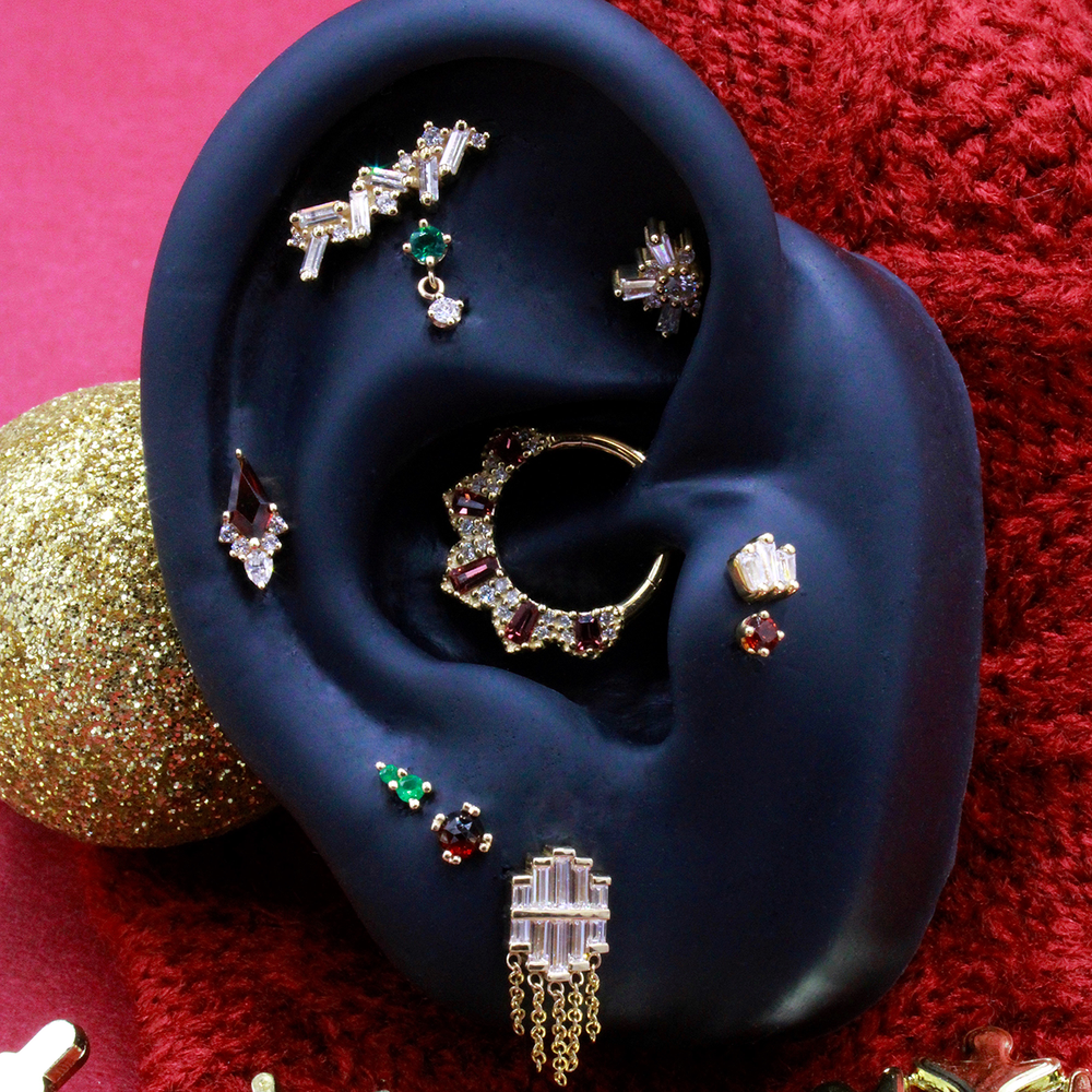 A curation of body jewelry on a black silicone ear display. This curation features the Garnet Afterglow in a helix along with a garnet prong in a tragus and other Buddha Jewelry and BVLA designs