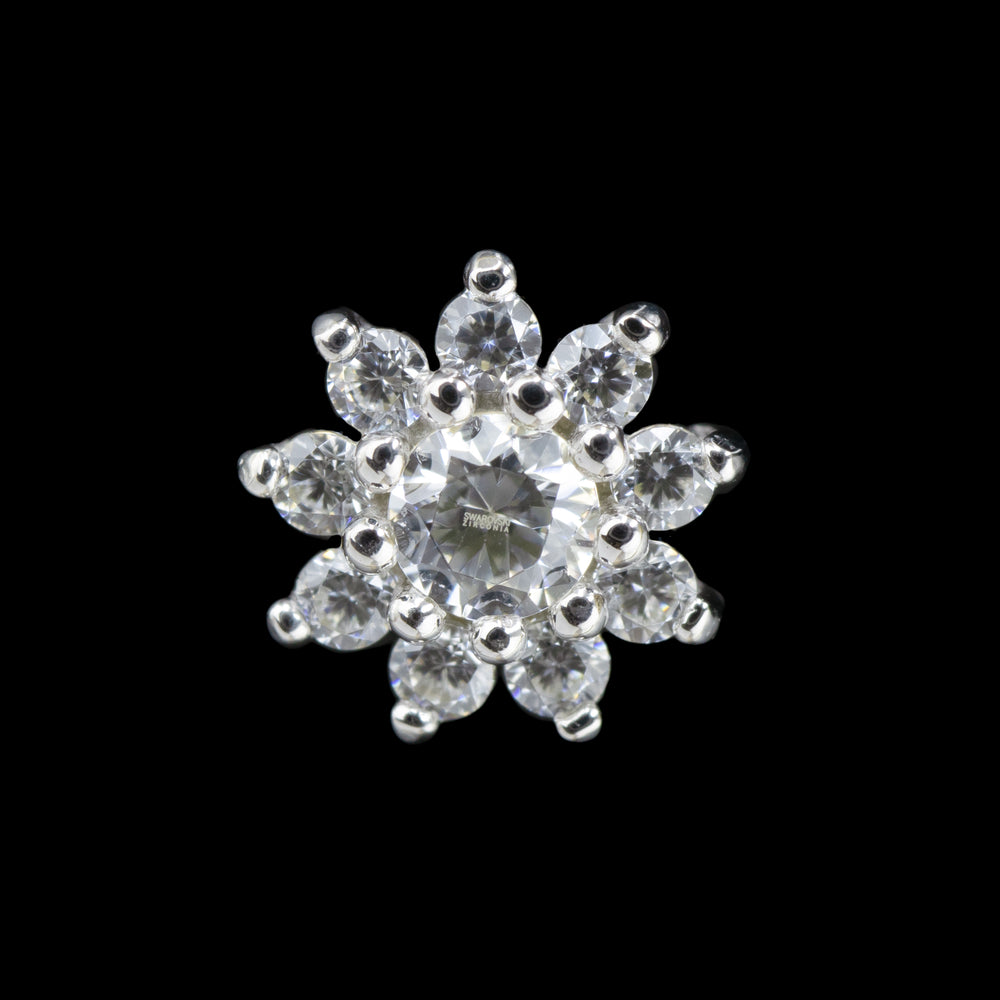 A white gold piece of body jewelry against a black background. The design is a similar shape to a daisy with a larger round cut cubic zirconia at it's center and 9 smaller round cut CZs surrounding the center gem. the gems are set in prongs. 