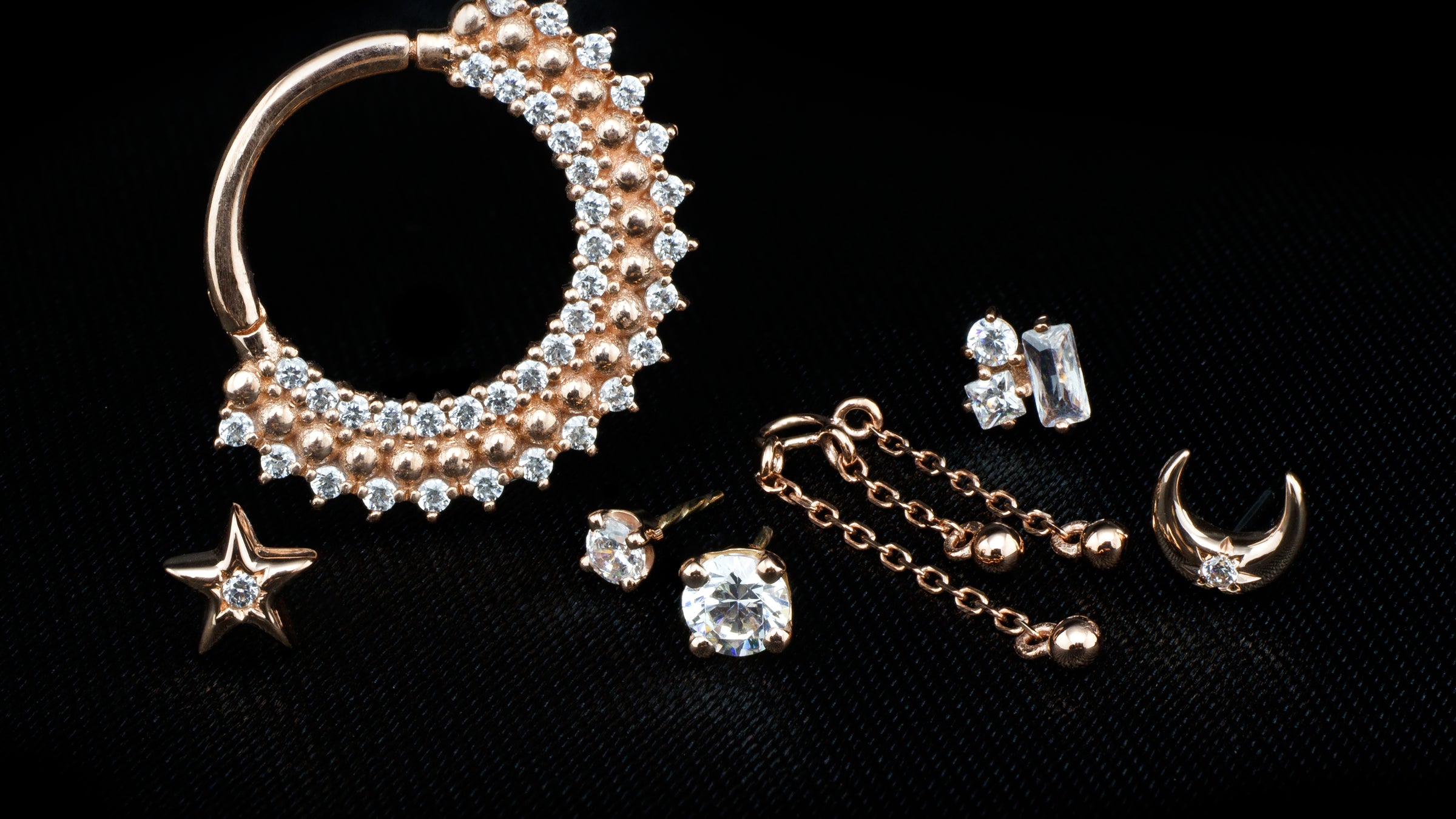 A collection of solid rose gold body jewelry pieces on a black cloth background. With white swarovski crystals
