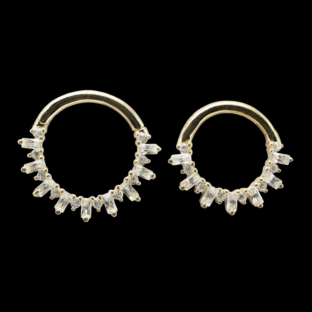 Two yellow gold pieces of body jewelry on a solid black background. The piece is a clicker hinge style ring with a high polish yellow gold wearing surface. The design of the ring consists of alternating round cut and baguette cut swarovski cubic zirconia. 