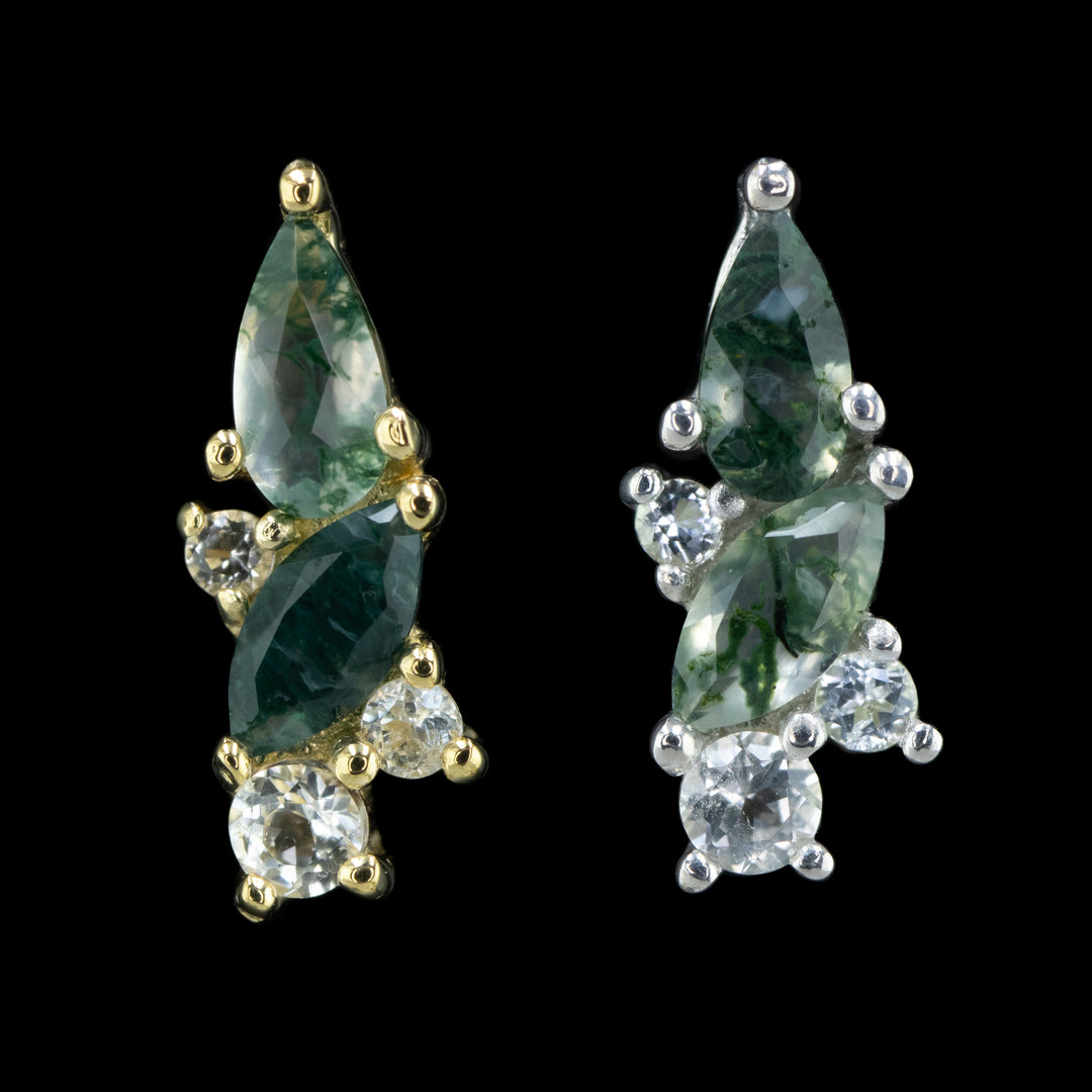 two pieces of body jewelry on a black background. The piece on the left is in yellow gold and the piece shown on the right id white gold. Both pieces feature a pear cut and a marquise cut moss agate and 3 ascending sizes of round cut white topaz. The design is slightly curved to the left.